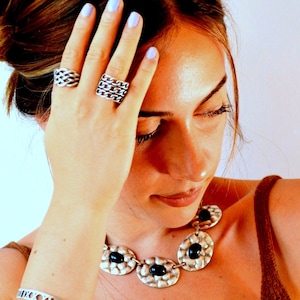 May include: Silver-coloured jewellery set featuring a necklace, rings, and bracelet. The necklace has oval pendants with black centre stones. The rings have chain-link designs. The bracelet has a repeating pattern. The jewellery is worn on a person.