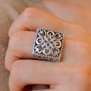 Op de afbeelding: Een zilverkleurige vierkante ring met een open ontwerp met een symmetrisch patroon. De ring wordt aan een vinger gedragen en toont de ingewikkelde details van het metaalwerk. Het ontwerp van de ring is een mix van geometrische vormen en bloemmotieven.