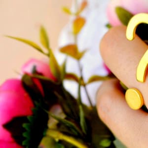 May include: A gold-coloured ring shaped like a question mark is worn on a finger. The ring has a curved top and a circular base. The background features pink and white flowers with green leaves.
