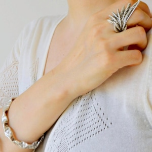 May include: A silver ring featuring a textured, multi-pronged design resembling a cluster of wheat or fern fronds, paired with a matching silver bangle bracelet with an irregular, hammered surface texture, worn on a white knit top.