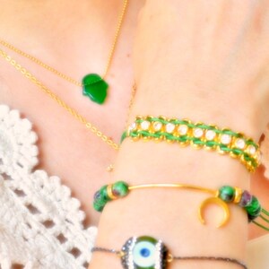 May include: A gold chain necklace with a green hamsa pendant, and three bracelets: one with green cord and clear stones, one with green and purple beads and a gold crescent moon charm, and one with a central green evil eye bead and black cord.
