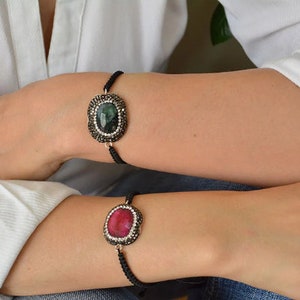 May include: Two bracelets featuring oval-shaped center stones, one dark green and one vibrant pink, each surrounded by a border of small, sparkling clear crystals. The stones are set in dark metal frames attached to thin, black woven cord bands.