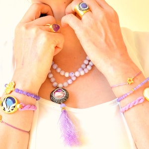 May include: An assortment of purple and gold jewelry. The image features a necklace with a large pendant and tassel, beaded bracelets, and gold rings with purple stones. The jewelry is set against a white background.