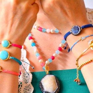 Könnte beinhalten: Eine Sammlung farbenfrohen Schmucks, darunter Armbänder mit Türkis- und rosa Steinen, ein blaues geflochtenes Armband mit einem großen Stein und eine Perlenkette mit Anhänger. Goldakzente sind sichtbar.