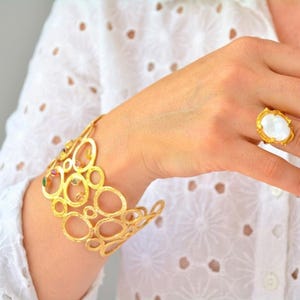 Può includere: Un bracciale a fascia dorato con un design traforato di cerchi interconnessi con piccole pietre preziose multicolori incastonate, abbinato a un anello dorato con una grande pietra bianca iridescente di forma ovale su una montatura testurizzata.
