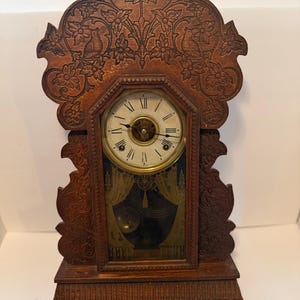 May include: A dark brown wooden mantel clock featuring intricate floral and bird carvings on the arched top. The clock face has black Roman numerals and two hands. Below the face, a glass panel reveals a gold-toned pendulum and curtain design.