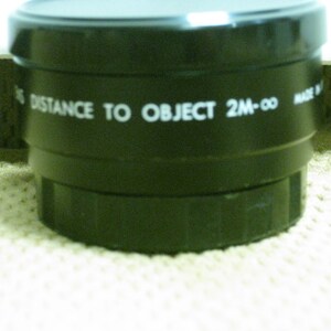 Puede incluir: Primer plano de una lente negra con texto blanco "DISTANCE TO OBJECT 2M-&infin;". La lente tiene un dise&ntilde;o estriado y est&aacute; sobre una superficie texturizada. Probablemente para una c&aacute;mara u otro dispositivo &oacute;ptico.