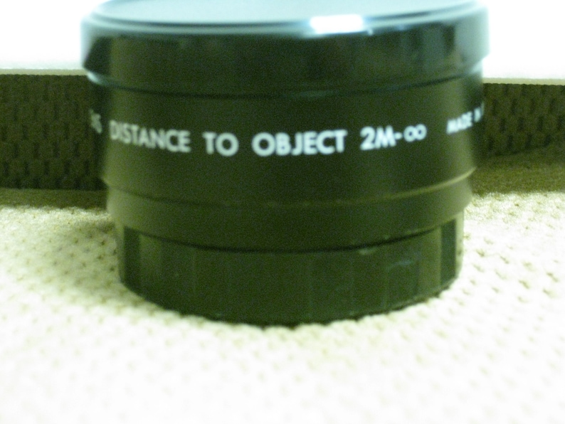 Puede incluir: Primer plano de una lente negra con texto blanco "DISTANCE TO OBJECT 2M-&infin;". La lente tiene un dise&ntilde;o estriado y est&aacute; sobre una superficie texturizada. Probablemente para una c&aacute;mara u otro dispositivo &oacute;ptico.