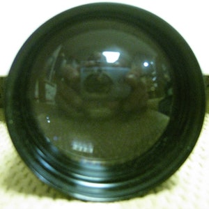 Puede incluir: Primer plano de una lente negra y convexa con una superficie reflectante. La lente est&aacute; rodeada por un marco negro. El reflejo muestra una imagen borrosa de una habitaci&oacute;n y una persona.