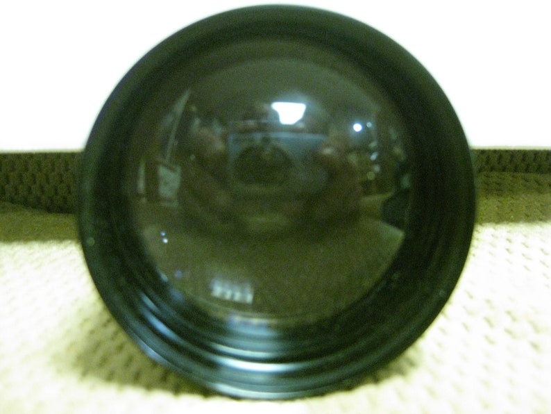 Puede incluir: Primer plano de una lente negra y convexa con una superficie reflectante. La lente est&aacute; rodeada por un marco negro. El reflejo muestra una imagen borrosa de una habitaci&oacute;n y una persona.