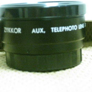 Puede incluir: Un objetivo negro ZYKKOR AUX. TELEPHOTO LENS I. La lente es cil&iacute;ndrica con un dise&ntilde;o estriado. El texto es blanco. La lente est&aacute; sobre una superficie blanca.