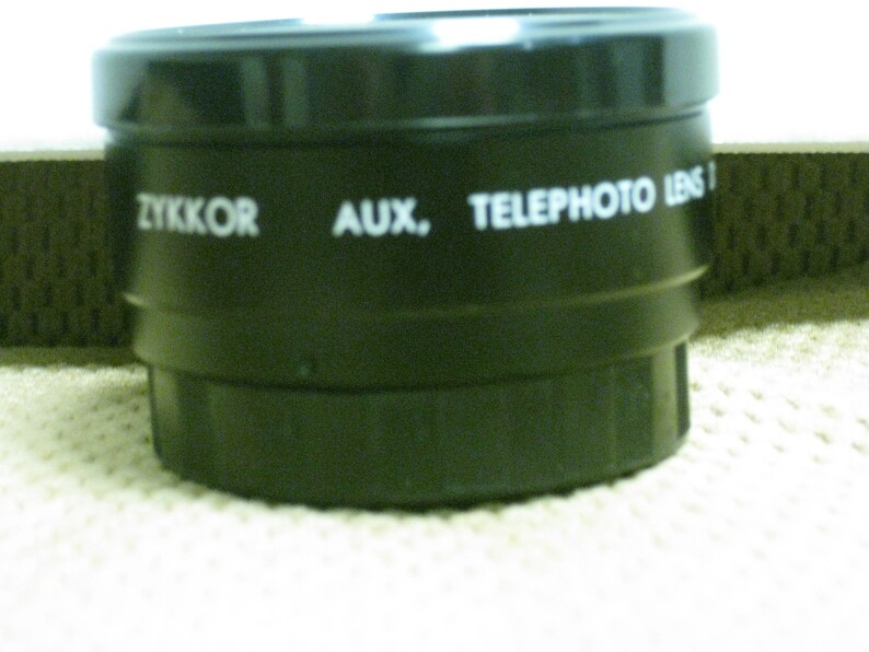 Puede incluir: Un objetivo negro ZYKKOR AUX. TELEPHOTO LENS I. La lente es cil&iacute;ndrica con un dise&ntilde;o estriado. El texto es blanco. La lente est&aacute; sobre una superficie blanca.