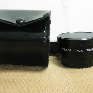 Puede incluir: Un objetivo ZYKKOR AUX. TELEPHOTO negro con un estuche de cuero negro a juego. La lente es cil&iacute;ndrica con texto blanco. El estuche es rectangular con cierre de bot&oacute;n a presi&oacute;n. Los art&iacute;culos est&aacute;n sobre un fondo neutro.