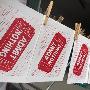May include: Five red and white printed tickets hanging on a clothesline with clothespins. Each ticket reads "ADMIT NOTHING" with the number "784496" printed on it.