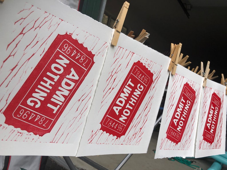 May include: Five red and white printed tickets hanging on a clothesline with clothespins. Each ticket reads "ADMIT NOTHING" with the number "784496" printed on it.