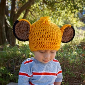 Puede incluir: Un niño con un gorro de león de crochet amarillo con orejas marrones. El gorro tiene un pequeño pompón marrón en la parte superior. El niño está sentado en una superficie de hormigón y lleva una camisa a rayas y pantalones cortos.