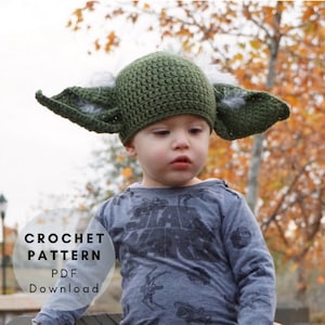 May include: A child wearing a green crocheted Yoda hat with white fur accents. The hat has large ears. The child is wearing a grey long-sleeved shirt with a black graphic print. The text "CROCHET PATTERN PDF DOWNLOAD" is visible in the image.