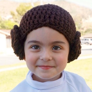 May include: A child wearing a brown crocheted hat with side buns. The hat is styled like Princess Leia's iconic hairstyle.