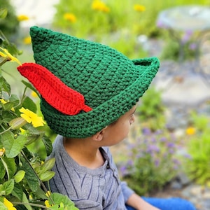 May include: A green crochet hat with a red feather accent. The hat has a pointed shape and is made of a textured yarn. The red feather is attached to the side of the hat. The hat is a costume accessory.
