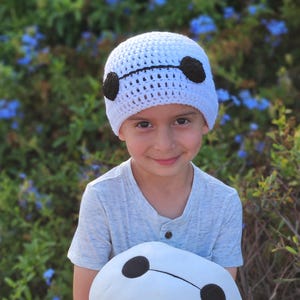 May include: A white crocheted beanie with black accents, resembling a character's design. The beanie is worn by a child, who is also holding a matching plush toy. The background features green foliage and blue flowers.