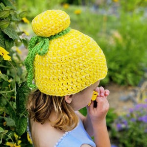 Op de afbeelding: Een handgemaakte gele gehaakte hoed met een groene stengel en een gele pompon bovenop. De hoed wordt door een persoon gedragen en de hoed is het middelpunt van de afbeelding. De hoed is gemaakt van zacht garen en is een uniek accessoire.