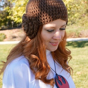 May include: A brown crocheted hat with a large flower detail on the side. The hat is worn by a person with long, red hair. The person is wearing a white shirt with a red and black pendant necklace featuring the Rebel Alliance symbol.