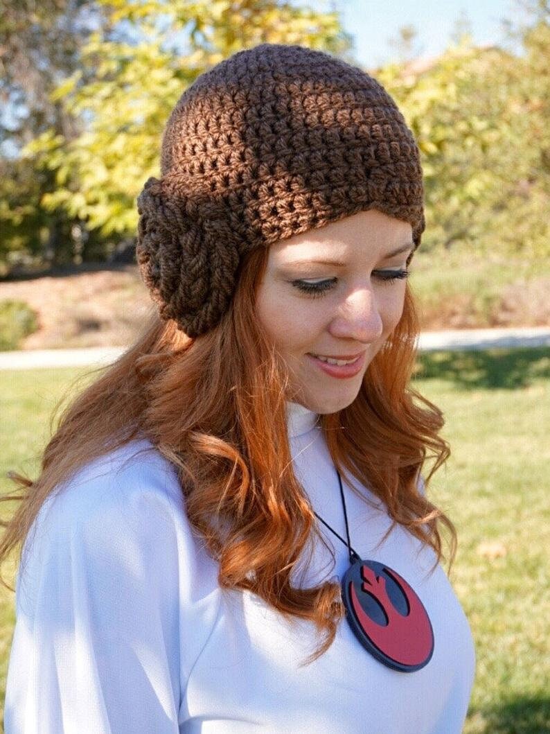 May include: A brown crocheted hat with a large flower detail on the side. The hat is worn by a person with long, red hair. The person is wearing a white shirt with a red and black pendant necklace featuring the Rebel Alliance symbol.