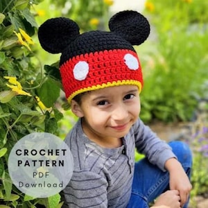 Peut inclure: Un bonnet crocheté à la main avec une base noire, deux grandes oreilles rondes, une section centrale rouge et une bordure jaune. Deux pièces circulaires blanches sont cousues sur le devant. Le texte CROCHET PATTERN PDF Download est visible.