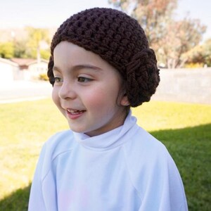 May include: A young person wearing a brown crocheted hat with a bun on the side, and a white turtleneck shirt.