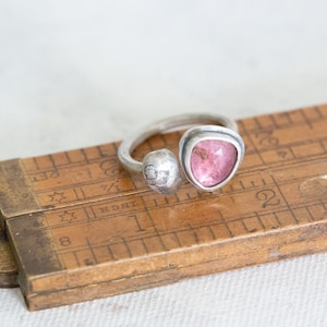Puede incluir: Un anillo de plata con una piedra preciosa rosa engastada en una banda retorcida. El anillo est&aacute; apoyado en una regla de madera.