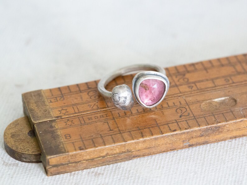 Puede incluir: Un anillo de plata con una piedra preciosa rosa engastada en una banda retorcida. El anillo est&aacute; apoyado en una regla de madera.