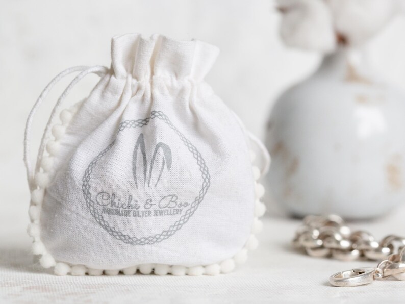 May include: A white cotton drawstring bag with a pom-pom trim. The bag has a gray logo that says "Chichi & Boo Handmade Silver Jewellery".