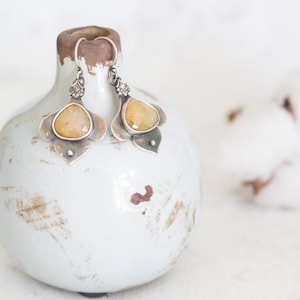 May include: A pair of silver earrings with yellow gemstone accents. The earrings are displayed on a white ceramic vase with a distressed finish.