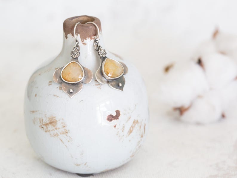 May include: A pair of silver earrings with yellow gemstone accents. The earrings are displayed on a white ceramic vase with a distressed finish.