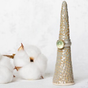 May include: A ring with a light green gemstone set in a silver band, displayed on a ceramic ring holder with a speckled brown and white glaze.