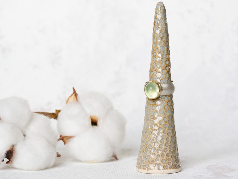 May include: A ring with a light green gemstone set in a silver band, displayed on a ceramic ring holder with a speckled brown and white glaze.