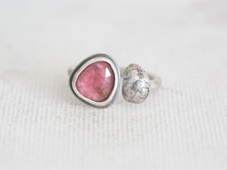 Puede incluir: Un anillo de plata con una piedra preciosa rosa engastada en un bisel triangular y una esfera de plata texturizada en la banda.
