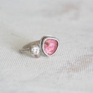 Puede incluir: Un anillo de plata con una piedra preciosa rosa engastada en forma triangular. El anillo tiene una banda de plata texturizada.