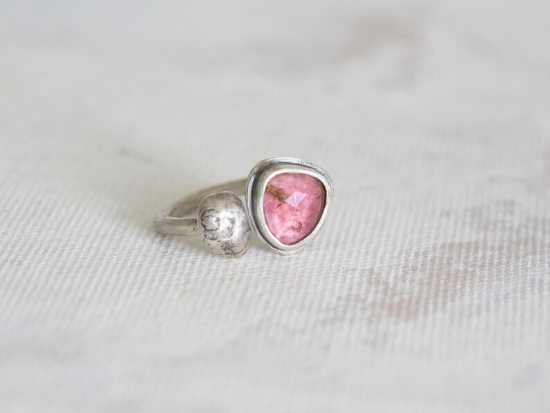 Puede incluir: Un anillo de plata con una piedra preciosa rosa engastada en forma triangular. El anillo tiene una banda de plata texturizada.
