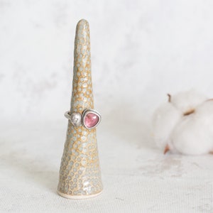Puede incluir: Un anillo de plata con una piedra preciosa rosa y blanca colocado en un soporte de anillo de cer&aacute;mica. El soporte del anillo est&aacute; salpicado de puntos blancos sobre un fondo marr&oacute;n claro.