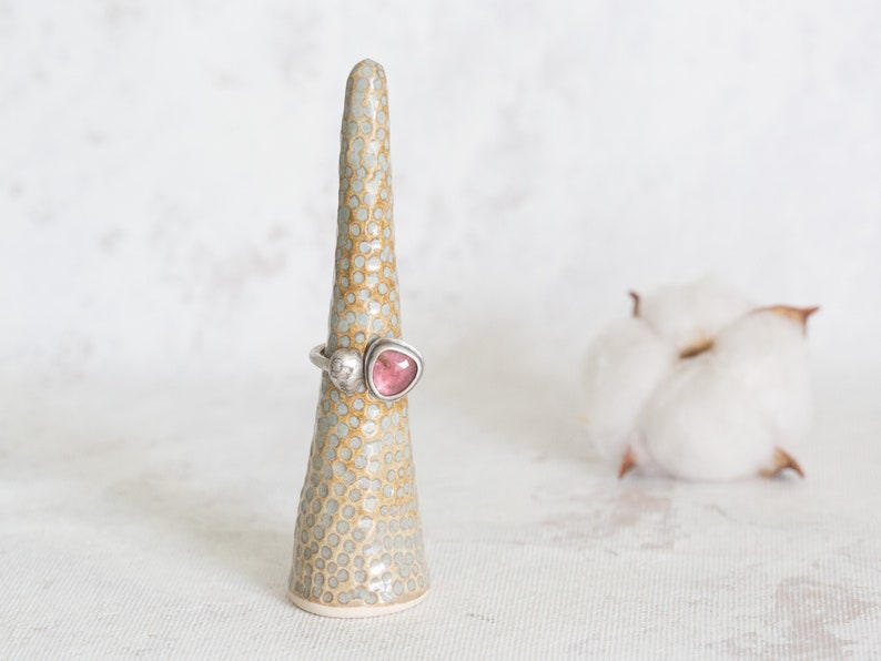 Puede incluir: Un anillo de plata con una piedra preciosa rosa y blanca colocado en un soporte de anillo de cer&aacute;mica. El soporte del anillo est&aacute; salpicado de puntos blancos sobre un fondo marr&oacute;n claro.
