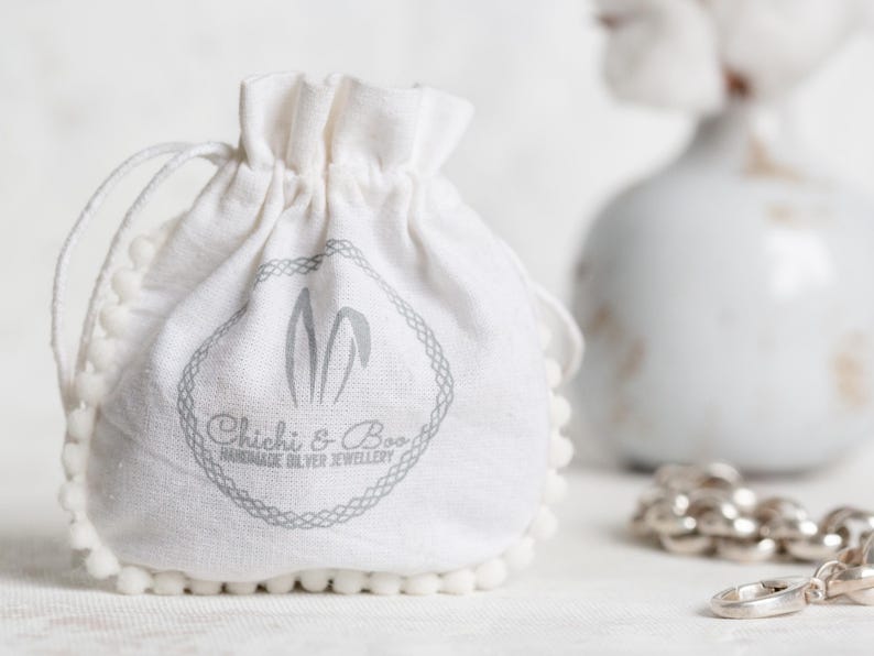 May include: A white cotton drawstring bag with a pom-pom trim. The bag has a gray logo that says "Chichi & Boo Handmade Silver Jewellery".