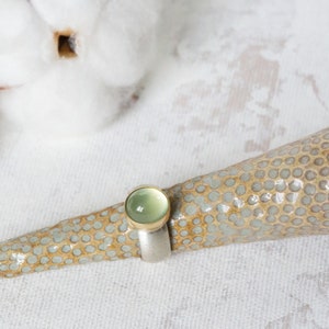 May include: A ring with a light green gemstone set in a gold band, displayed on a ceramic ring holder with a speckled pattern.