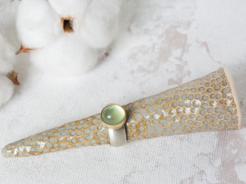 May include: A ring with a light green gemstone set in a gold band, displayed on a ceramic ring holder with a speckled pattern.