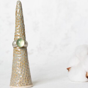 May include: A silver ring with a green gemstone set in a gold band. The ring is displayed on a ceramic ring holder.