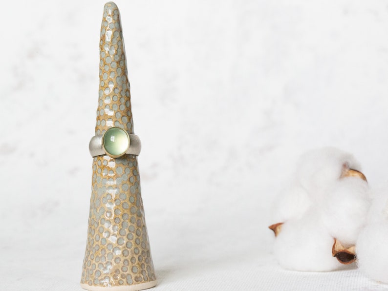 May include: A silver ring with a green gemstone set in a gold band. The ring is displayed on a ceramic ring holder.
