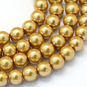 May include: A strand of gold-colored round pearl beads. The beads are smooth and shiny, and they are arranged in a loose pile.