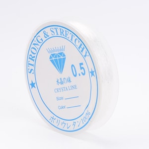 May include: A white spool of strong and stretchy elastic cord with a blue label that reads "STRONG & STRETCHY", "水晶の話", "CRYSTA LINE", "0.5", "Size:", "Color:", and "ボリウレタン100%".