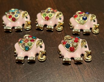 May include: Five pink elephant-shaped brooches with gold-colored accents. Each elephant is adorned with colorful, sparkling gemstones on its back. The brooches are displayed on a dark brown surface.