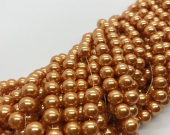 May include: A pile of gold-colored round pearl beads. The beads are arranged in a circular pattern, with the beads in the center of the circle being the largest.